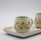 Two ceramic mugs with leaf patterns on a white tray against a light gray background