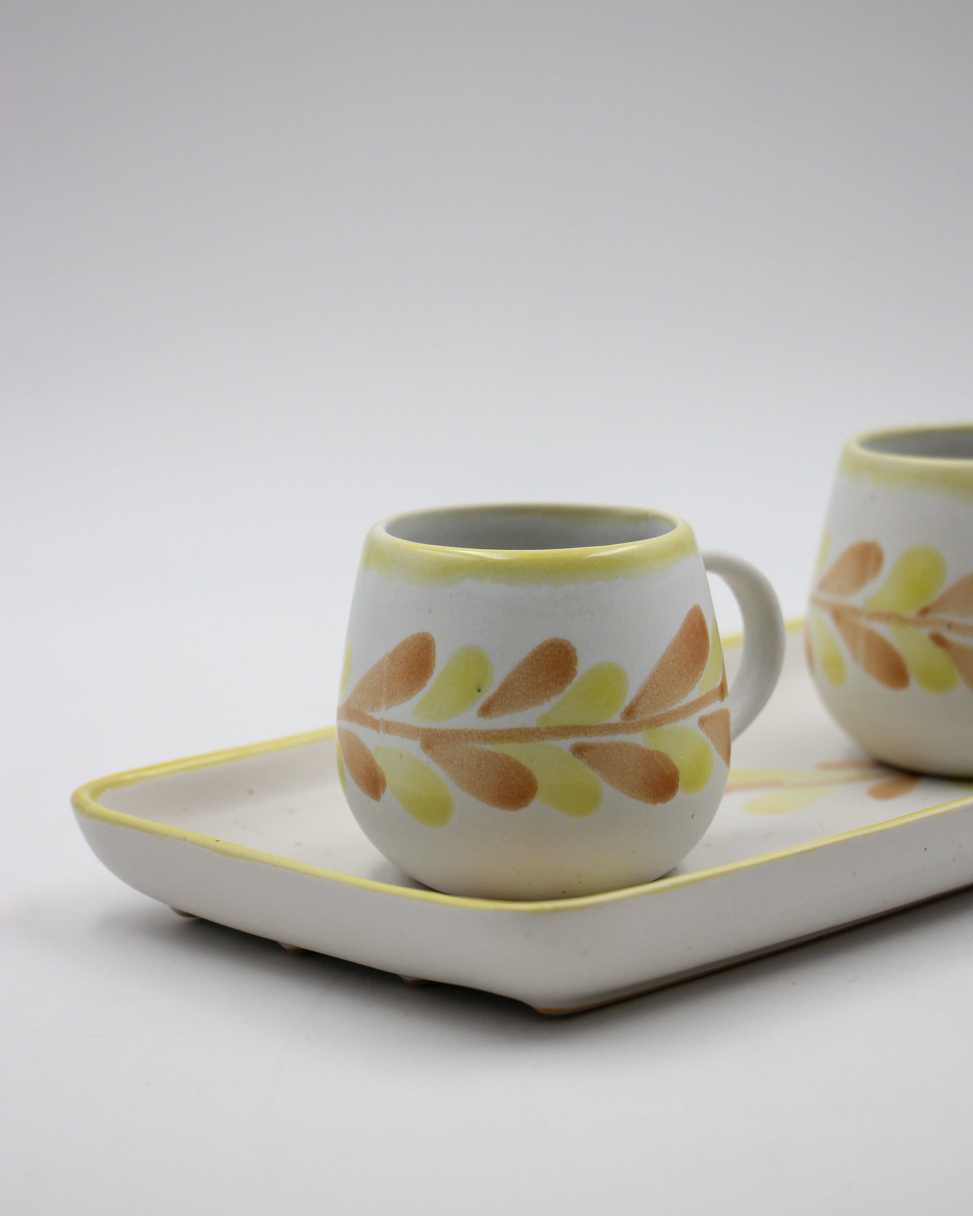 Two ceramic mugs with leaf patterns on a white tray against a light gray background