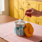 Person scooping something from a floral-patterned container with a yellow lid in a kitchen setting.